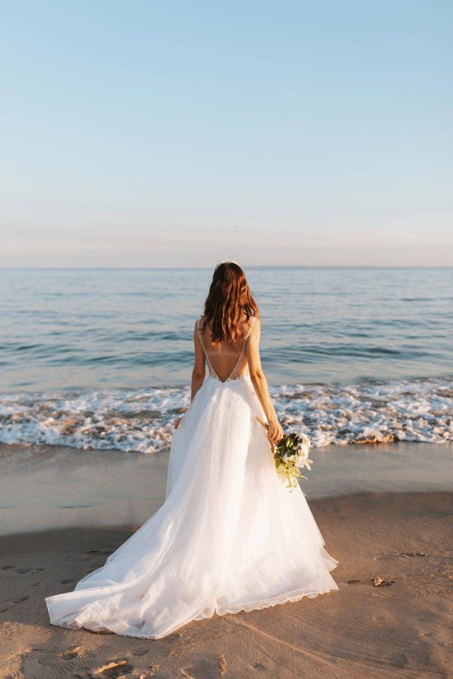 Bride in white wedding dress with bouquet stands on a beach in Key West and looks out at water