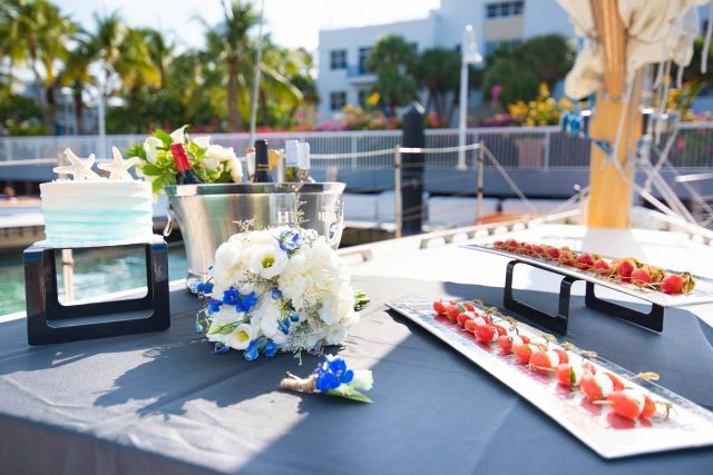 Setup for a sailboat wedding aboard a schooner with a bouquet of flowers, a cake, appetizers and chilled wine
